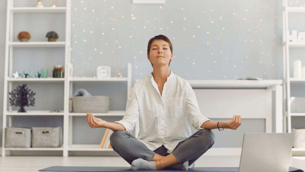 a woman meditating on a yoga mat in front of a laptop